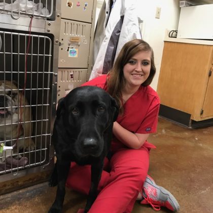 A team member in red scrubs sitting on the floor cuddling a black lab